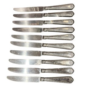 May include: A collection of nine silver-colored butter knives. Each knife has a rounded blade and a decorative handle with a floral design. The knives are arranged vertically against a white background.