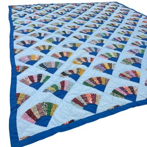 May include: A quilt featuring a repeating fan pattern made of various multi-colored floral fabric scraps. The fan shapes are set against a light blue background with a solid dark blue border framing the entire rectangular textile piece.
