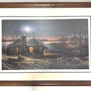 May include: A framed print of a painting depicting a cozy cabin in the woods under a starry night sky. The cabin has a stone exterior and a warm glow emanating from the windows. A dog is lying in the grass in the foreground. The title of the painting is "First Christmas" and is visible at the bottom of the print.