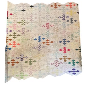 Puede incluir: Una colcha rectangular con una base de color crema y un patrón de cuadrícula de diamantes repetitivo. Dispersos por toda la cuadrícula hay pequeños grupos de diamantes bordados multicolores en tonos rojo, azul, verde, amarillo, morado y naranja.