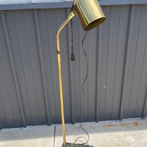 Puede incluir: Una lámpara de pie vintage de latón con una pantalla cilíndrica y un brazo delgado y curvo. La lámpara tiene un acabado dorado y se apoya sobre una base cuadrada negra. Un cable eléctrico marrón se extiende desde la lámpara.