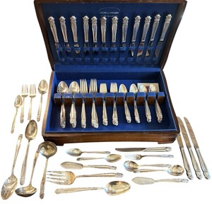 May include: A complete set of silver silverware, including forks, spoons, knives, and serving utensils, displayed in a wooden case with a blue velvet interior. The silverware has ornate floral detailing.