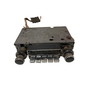 May include: Vintage car radio with a metal faceplate and chrome knobs. The radio has AM and FM buttons, a tuning dial, and a series of buttons. Wires extend from the top of the radio. The radio is designed to fit into a car dashboard.