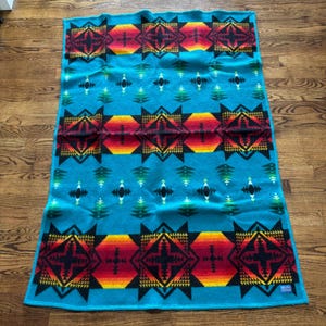 Puede incluir: Una manta rectangular con un fondo turquesa vibrante y tres bandas horizontales de patrones geométricos en negro, rojo, naranja y amarillo. Pequeños motivos en forma de diamante negros y verdes se intercalan entre las bandas.