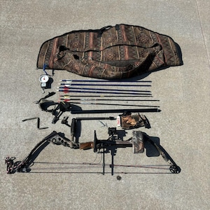 May include: A black compound bow with a camouflage riser and limbs, a black arrow rest, and a black sight. The bow is laid out on a gray concrete surface with a quiver of arrows, a bow stringer, a bow glove, and a bow case.