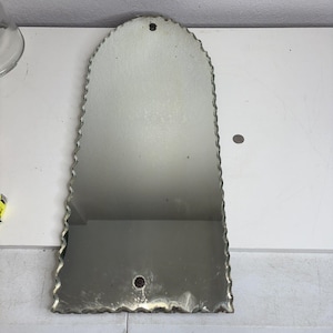 May include: A vintage, arched mirror with a scalloped edge. The mirror has a silvered surface and a small hole at the top for hanging. A decorative element is at the bottom. The mirror is a decorative item.