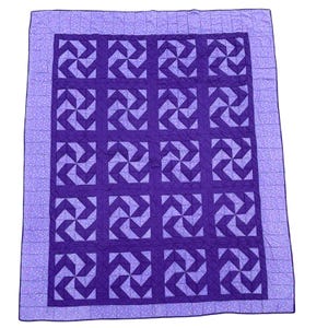 Op de afbeelding: Een handgemaakte quilt met een windmolenontwerp in paarse tinten. De quilt heeft een donkerpaars midden met lichtere paarse windmolenblokken. Een lichtpaarse rand met een bloemenpatroon omlijst de quilt.