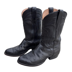 May include: A pair of black leather cowboy boots featuring a textured lizard-skin pattern on the foot and a smooth shaft with decorative stitching. The boots have a pointed toe, a low stacked heel, and pull tabs at the top of the shaft.