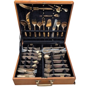 May include: A vintage wooden cutlery case with a brass handle and hinges, open to reveal a complete set of gold-colored flatware. The set includes knives, forks, spoons, and serving utensils, all neatly arranged on a black velvet interior.