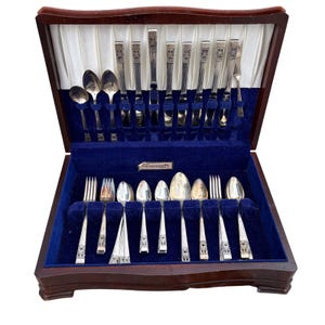 May include: A wooden silverware chest with a dark finish, lined with blue velvet. The chest contains a complete set of silver-colored flatware, including forks, spoons, and knives, arranged neatly within the compartments. The set is likely vintage.