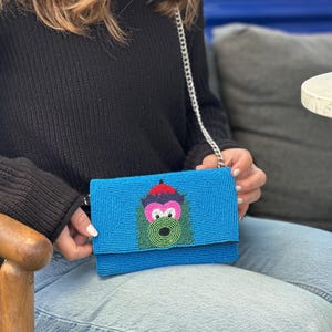 Può includere: Una pochette rettangolare in perline blu con un design grafico centrale raffigurante una creatura stilizzata con cappello rosso, occhi rosa e viso verde. La borsa include una tracolla a catena in metallo argentato e una chiusura a patta.