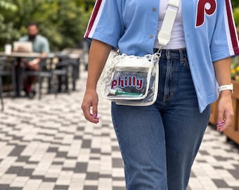 May include: A clear plastic crossbody bag with white trim and a white strap. The front of the bag features the word Philly in red and blue lettering. The bag is worn over a light blue button-down jersey with red and white sleeve stripes.