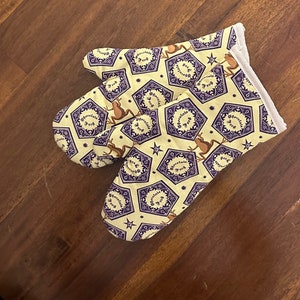 May include: A pair of yellow oven mitts with a purple and white pattern featuring a frog and the text "Chocolate Frog".