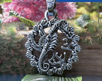 May include: Silver pendant featuring a detailed, ornate design of a seahorse with a circular frame of stylized shells. The pendant is on a wooden surface with green foliage in the background.