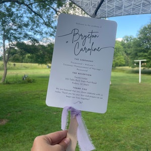 May include: White wedding fan with a purple ribbon. The fan has the text "Welcome to the wedding of Brycton + Caroline" and the ceremony and reception details.