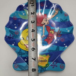 May include: A shell-shaped plate featuring Ariel, Flounder, and Sebastian from The Little Mermaid. The plate is blue with underwater graphics and a Disney's The Little Mermaid logo. A metal measuring tape shows the plate is 8 inches in length.