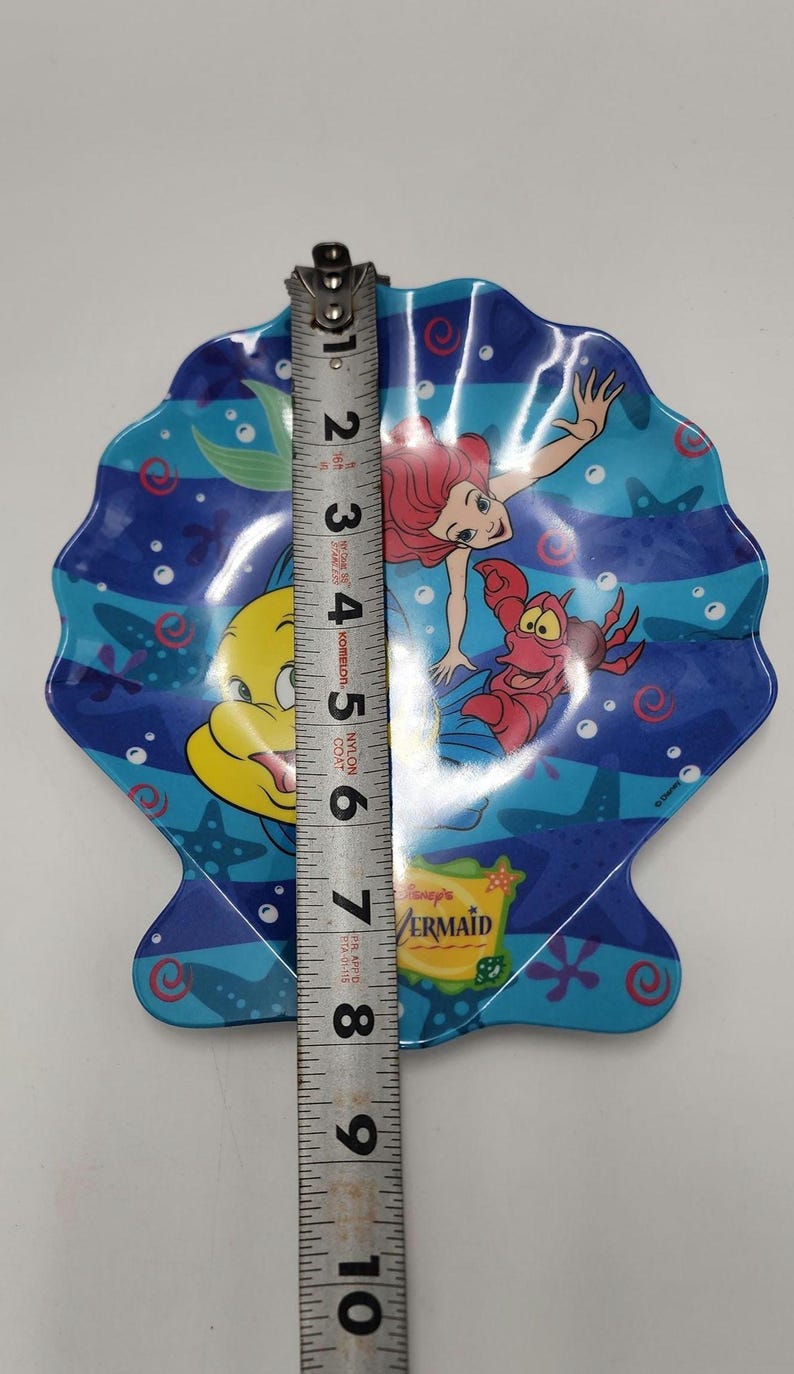 May include: A shell-shaped plate featuring Ariel, Flounder, and Sebastian from The Little Mermaid. The plate is blue with underwater graphics and a Disney's The Little Mermaid logo. A metal measuring tape shows the plate is 8 inches in length.