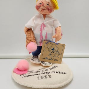 May include: A felt doll wearing a yellow cap, white shirt with pink stains, and blue trousers, holding a waffle cone with a pink pom-pom ice cream scoop. The doll stands on a circular base labelled Annalee Doll Society Anniversary Edition 1993.