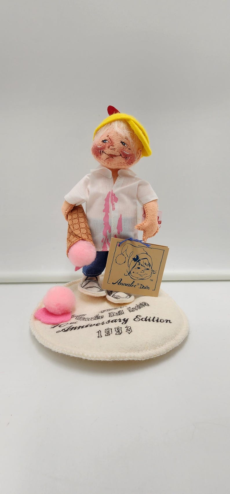 May include: A felt doll wearing a yellow cap, white shirt with pink stains, and blue trousers, holding a waffle cone with a pink pom-pom ice cream scoop. The doll stands on a circular base labelled Annalee Doll Society Anniversary Edition 1993.