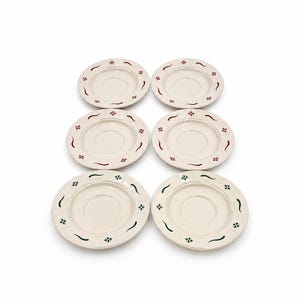 May include: A set of six round ceramic saucers in an off-white colour. Each saucer features a raised rim decorated with a repeating pattern of small floral motifs and curved lines in alternating red and dark green colours around the edge.