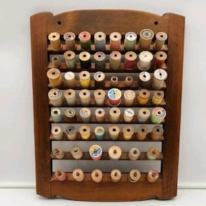 May include: A rectangular wooden wall-mounted rack featuring multiple horizontal slats designed to hold numerous vintage sewing thread spools. The spools are arranged in rows and display a variety of colors, including blue, yellow, pink, and white.