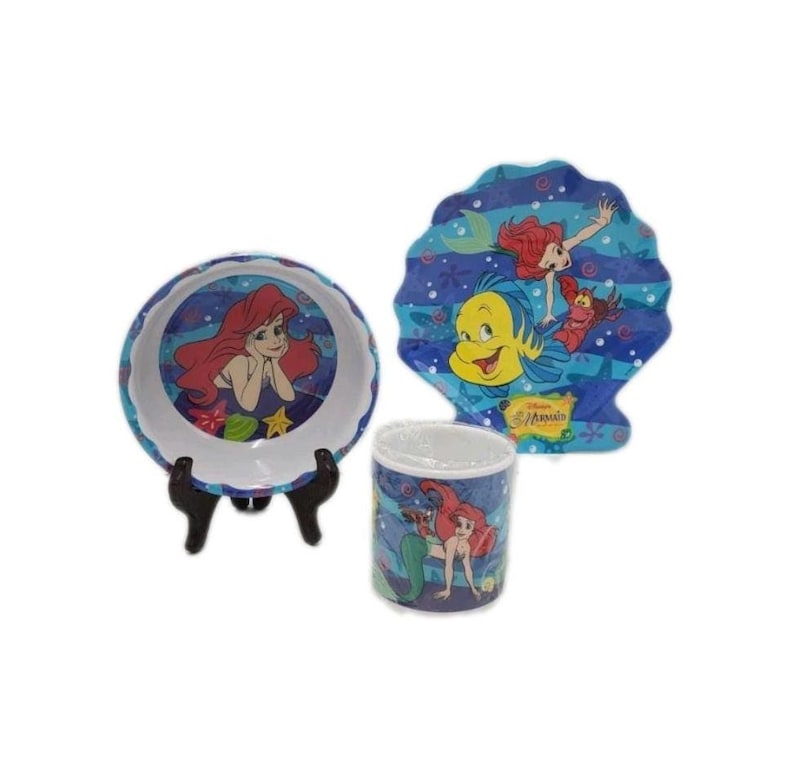 May include: A Disney The Little Mermaid dining set featuring a round bowl, a cylindrical mug, and a shell-shaped plate. Each piece displays colorful illustrations of Ariel, Flounder, and Sebastian against a blue underwater background with bubbles.