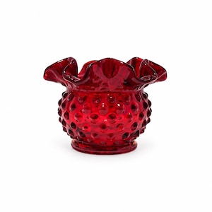 May include: A deep red glass bowl featuring a hobnail texture across its rounded body and a wide, ruffled, crimped rim. The translucent glass vessel sits on a circular base, showcasing a vibrant crimson hue and a distinctively patterned surface.