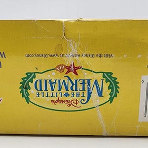 May include: A yellow rectangular cardboard box for a Disney The Little Mermaid product. The box features the logo, a barcode, and text including Nontoxic, Dishwasher Safe, Wash Before Using, and Zak Designs Inc. branding with a copyright symbol.