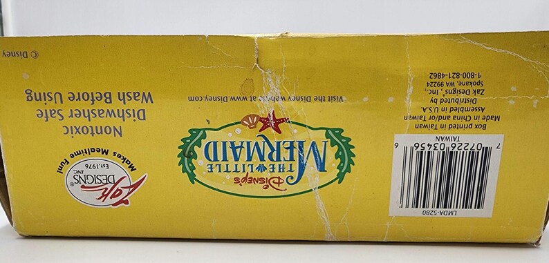 May include: A yellow rectangular cardboard box for a Disney The Little Mermaid product. The box features the logo, a barcode, and text including Nontoxic, Dishwasher Safe, Wash Before Using, and Zak Designs Inc. branding with a copyright symbol.