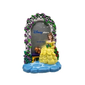 May include: A Disney Store picture frame featuring Belle from Beauty and the Beast. The frame is white with a floral arch, purple flowers, and a blue base. Belle is in a yellow dress, and a squirrel is seated on a bench.