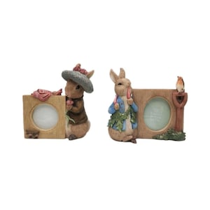 Puede incluir: Dos marcos de fotos decorativos con personajes de Peter Rabbit. Un marco tiene un conejo sosteniendo una flor, el otro tiene un conejo sosteniendo una zanahoria y un pájaro. Ambos marcos son de madera y tienen aberturas circulares.