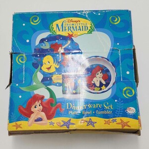 May include: A Disney The Little Mermaid dinnerware set box featuring Ariel and Flounder. The blue packaging displays a plate, bowl, and tumbler with underwater graphics, yellow starfish, and the text Disney's The Little Mermaid Dinnerware Set.