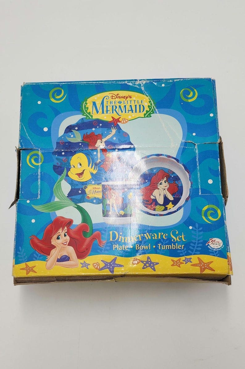 May include: A Disney The Little Mermaid dinnerware set box featuring Ariel and Flounder. The blue packaging displays a plate, bowl, and tumbler with underwater graphics, yellow starfish, and the text Disney's The Little Mermaid Dinnerware Set.