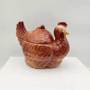 May include: A ceramic chicken-shaped covered dish in shades of brown, with a red comb and wattle. The dish is designed to resemble a hen, with detailed feather textures and a curved shape. The lid fits snugly, creating a functional and decorative kitchen item.