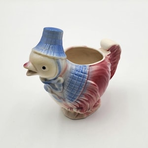 May include: A vintage ceramic creamer shaped like a duck, featuring a blue hat, a blue and white bib, and a red and white body. The creamer has a handle and a spout for pouring.