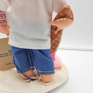 May include: A doll wearing a white shirt and blue trousers, holding a brown textured object. A white tag attached to the arm reads Annalee '93 MADE ONLY IN MEREDITH NEW HAMPSHIRE U.S.A. A tan card stands behind the doll on a circular white base.