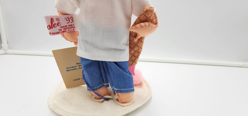 May include: A doll wearing a white shirt and blue trousers, holding a brown textured object. A white tag attached to the arm reads Annalee '93 MADE ONLY IN MEREDITH NEW HAMPSHIRE U.S.A. A tan card stands behind the doll on a circular white base.