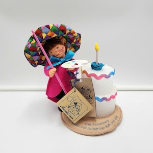 May include: A felt doll wearing a bright pink coat and a multi-coloured diamond-patterned umbrella, holding a painter's palette. Beside the doll is a two-tier white cake with a single candle. A tan tag reads Annalee Doll 15th Anniversary Logo Kid.
