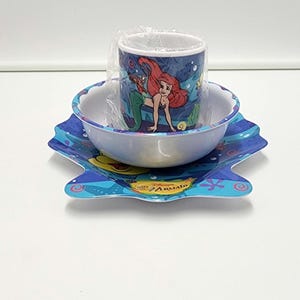 May include: A three-piece dining set featuring a blue plastic plate, a white bowl, and a mug wrapped in clear plastic. The items are decorated with illustrations of a red-haired mermaid and sea life, with the text Disney The Little Mermaid.