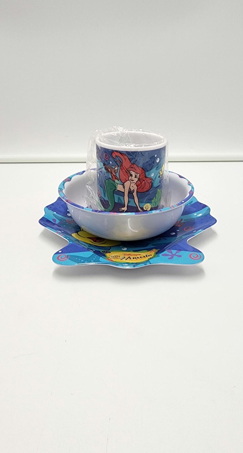 May include: A three-piece dining set featuring a blue plastic plate, a white bowl, and a mug wrapped in clear plastic. The items are decorated with illustrations of a red-haired mermaid and sea life, with the text Disney The Little Mermaid.
