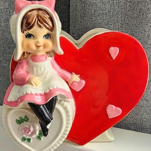 May include: A vintage ceramic Valentine's Day figurine. It features a smiling girl in a pink dress and bonnet, seated on a heart-shaped base with a rose. A large red heart with pink hearts is behind her.