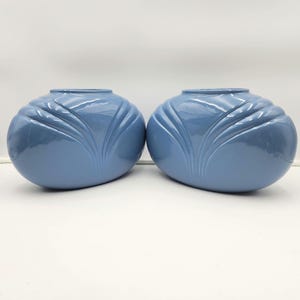 Puede incluir: Dos jarrones de cerámica azules a juego. Cada jarrón tiene una forma redondeada con una serie de crestas verticales curvas. Los jarrones son de un color azul liso y apagado y tienen un acabado brillante. Se exhiben sobre una superficie blanca.