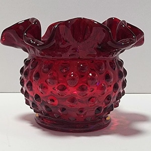 May include: A ruby red glass hobnail bowl with a ruffled rim. The bowl has a round shape and a hobnail pattern covering the exterior. The rim is wavy and adds a decorative touch to the piece. The bowl sits on a small, round base.