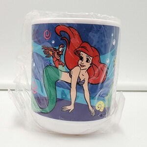 May include: A white ceramic mug wrapped in clear plastic film featuring a colorful graphic of a red-haired mermaid with a green tail and a small crab character. The background of the design is a deep blue ocean scene with bubbles and sea shells.