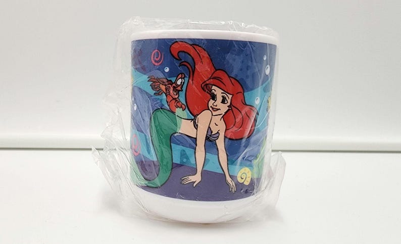 May include: A white ceramic mug wrapped in clear plastic film featuring a colorful graphic of a red-haired mermaid with a green tail and a small crab character. The background of the design is a deep blue ocean scene with bubbles and sea shells.