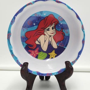 May include: A round white bowl with a scalloped blue rim featuring a pattern of red and blue swirls. The center of the bowl displays a colorful illustration of a red-haired character resting their chin on their hands, surrounded by a pink starfish, a green seashell, and a yellow starfish against a blue water background.