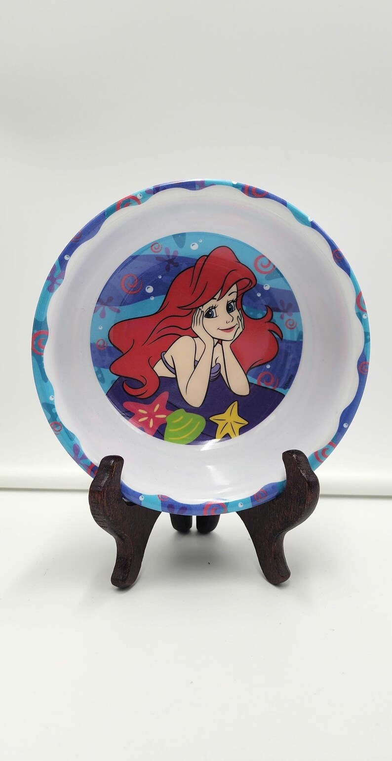 May include: A round white bowl with a scalloped blue rim featuring a pattern of red and blue swirls. The center of the bowl displays a colorful illustration of a red-haired character resting their chin on their hands, surrounded by a pink starfish, a green seashell, and a yellow starfish against a blue water background.