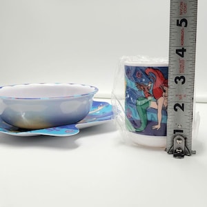 May include: A children's bowl and cup set featuring Ariel and Flounder graphics. The cup is wrapped in clear plastic and stands approximately 3.5 inches tall next to a metal measuring tape. The bowl has a scalloped edge and blue base.