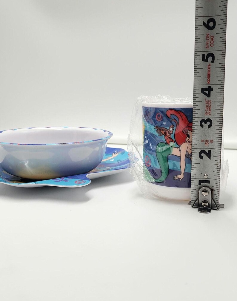 May include: A children's bowl and cup set featuring Ariel and Flounder graphics. The cup is wrapped in clear plastic and stands approximately 3.5 inches tall next to a metal measuring tape. The bowl has a scalloped edge and blue base.