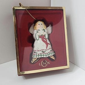 May include: A porcelain angel Christmas ornament in a red velvet box with a gold trim. The angel is wearing a white dress with red and green trim and holding a star. The box has the word "LENOX" printed on it.